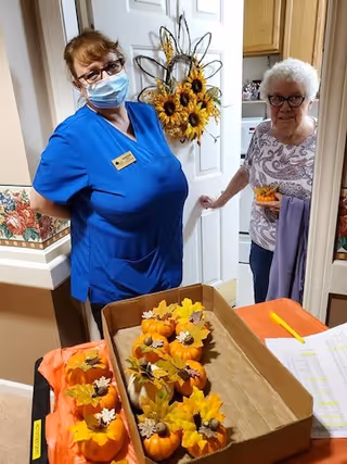 A staff member wearing blue scrubs and a face mask stands next to an elderly woman with white hair and glasses inside a retirement facility. They are near a table with a box containing small decorative pumpkins and autumn leaves. A sunflower wreath hangs on the door behind them.
