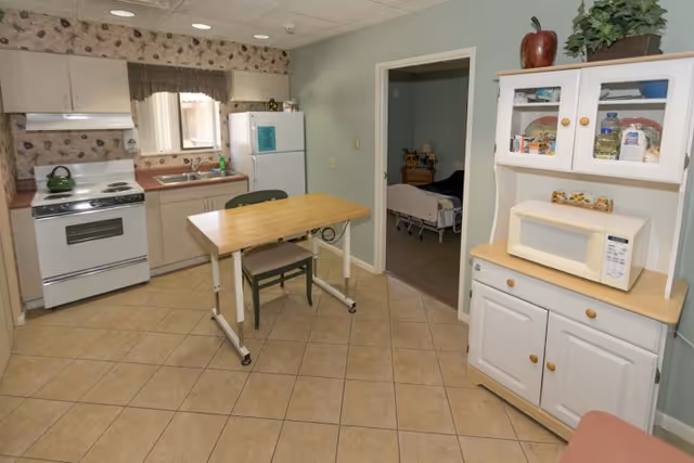 A small kitchen area with a stove, refrigerator, sink, and cabinets. There is a wooden table with a chair in the center of the tiled floor. A white hutch with a microwave and various items is on the right side. A doorway leads to a bedroom with a hospital bed visible.