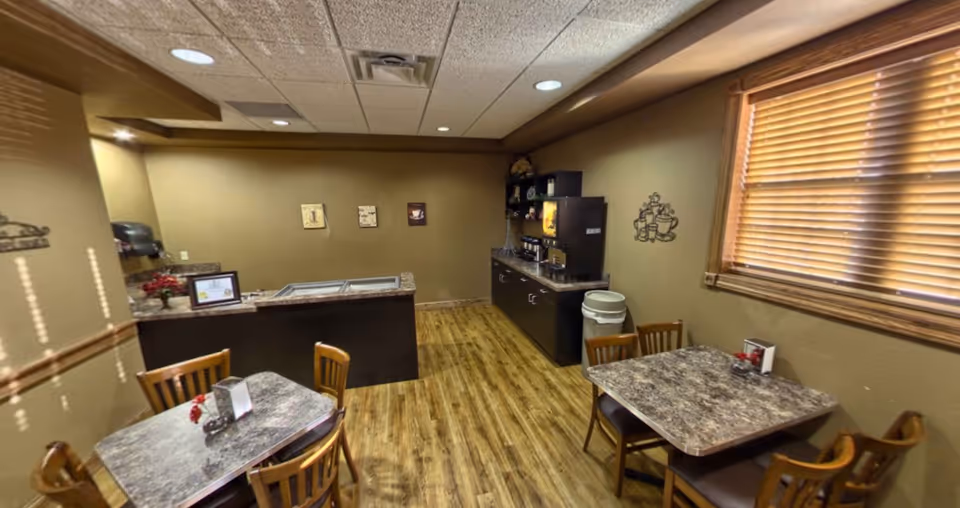 Small dining area with two square tables, each surrounded by four wooden chairs. The room has wood flooring, beige walls, and a window with wooden blinds. There is a counter with a coffee machine and other appliances along one wall, and a trash can nearby.
