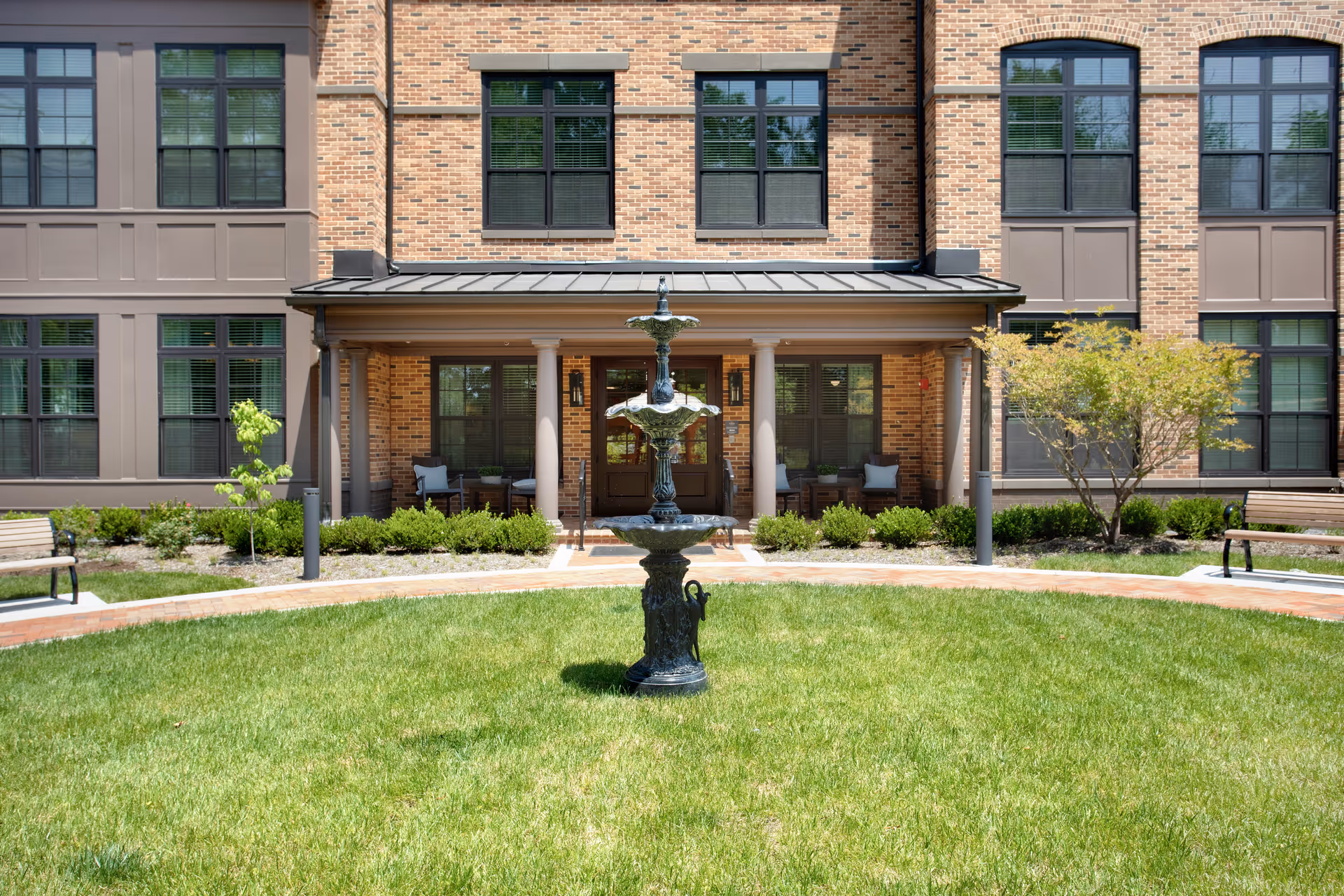 Outdoor courtyard area of a senior living facility with a central decorative fountain, green grass, benches on either side, and a brick building with large windows and a covered porch with seating in the background.