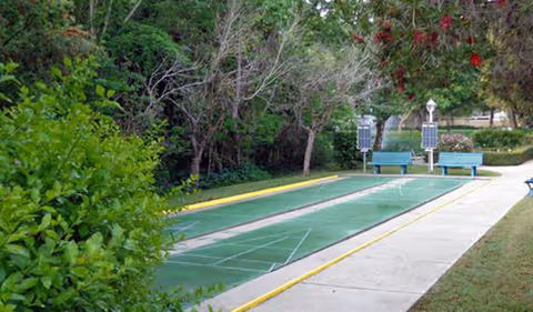 Outdoor bocce ball court with two benches and solar-powered lights along a paved walkway, surrounded by trees and greenery.