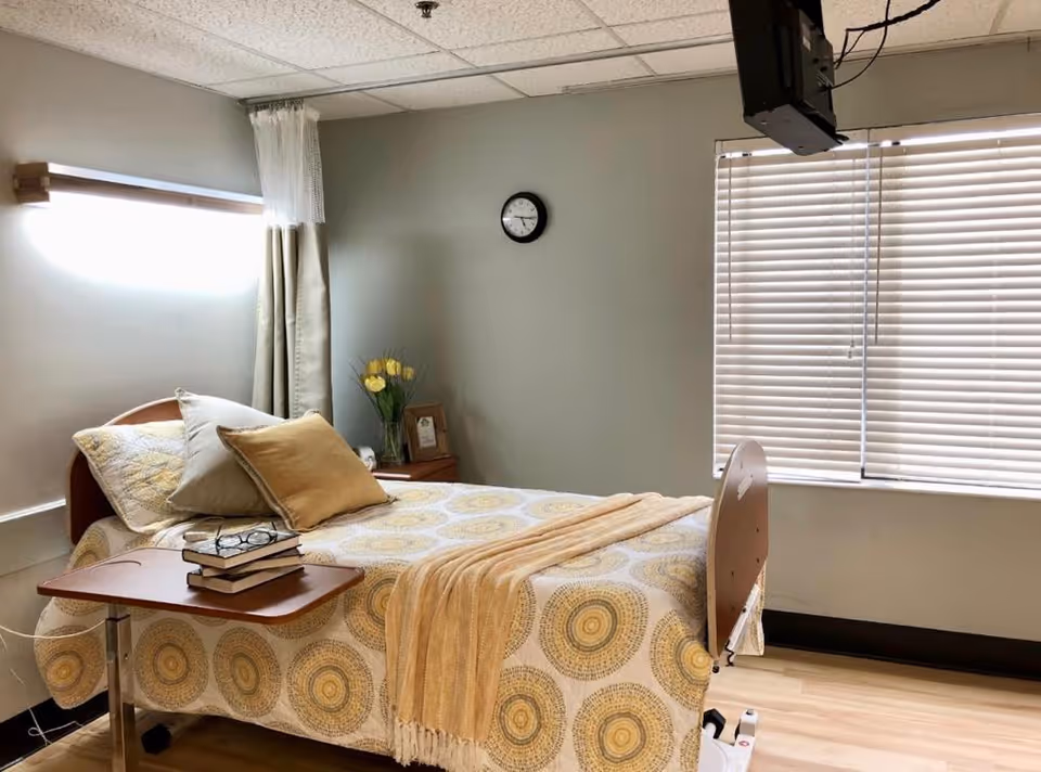 A tidy senior-care bedroom with a single bed, bedside table with flowers and books, window blinds, and a wall-mounted TV.