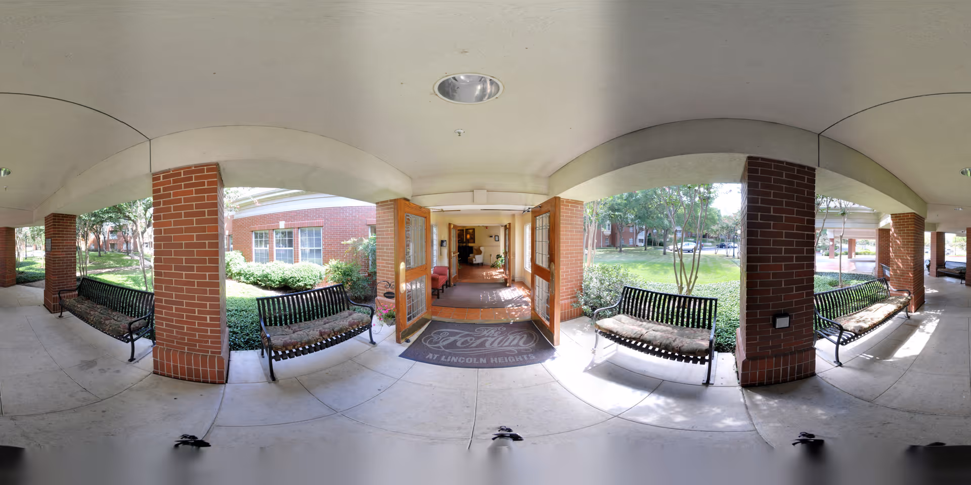 Covered outdoor walkway with brick pillars and black metal benches with cushions on either side. The entrance to a building is visible with double wooden doors open, revealing an interior hallway. Green bushes and trees surround the walkway and building exterior.