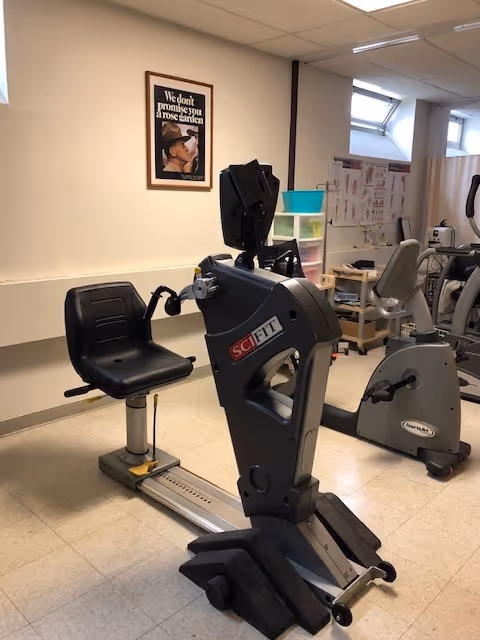 Indoor exercise room with a seated exercise bike labeled SCI FIT in the foreground and another exercise bike in the background. The room has light-colored walls and flooring, a framed motivational poster on the wall, and storage shelves with various items. There are anatomical charts on the wall near a window.