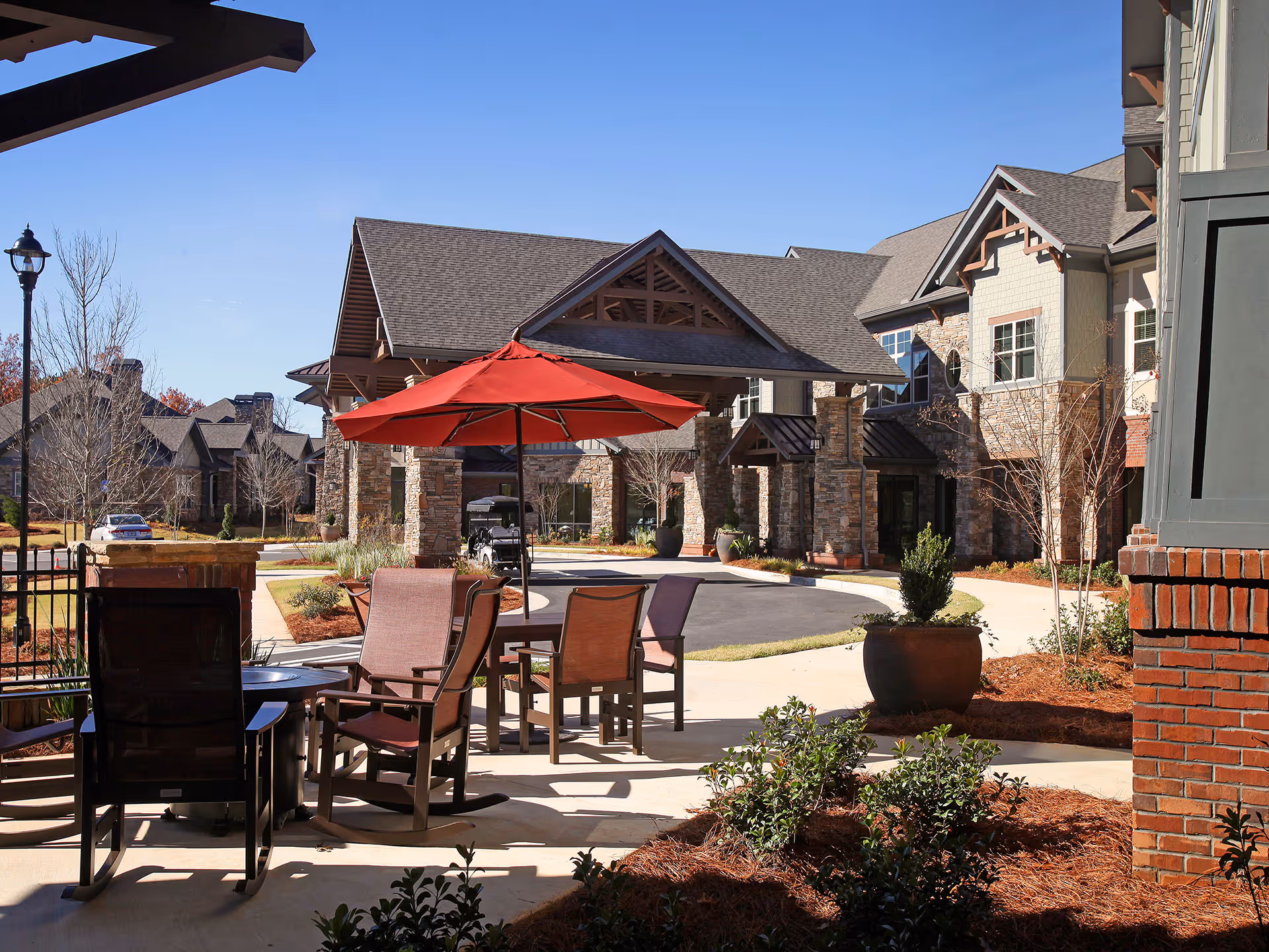 Outdoor seating area with a red umbrella and chairs in front of the entrance to a multi-story senior living building.