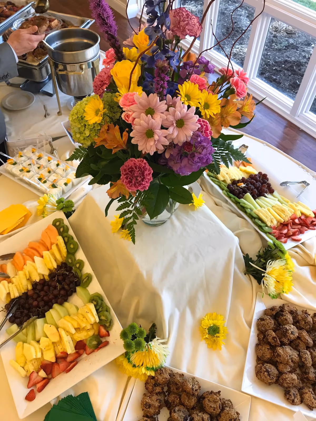 A buffet table with a large colorful floral arrangement in the center, surrounded by platters of assorted fresh fruit, cookies, and deviled eggs. The table is covered with a white tablecloth and is set near large windows with a view of the outdoors.