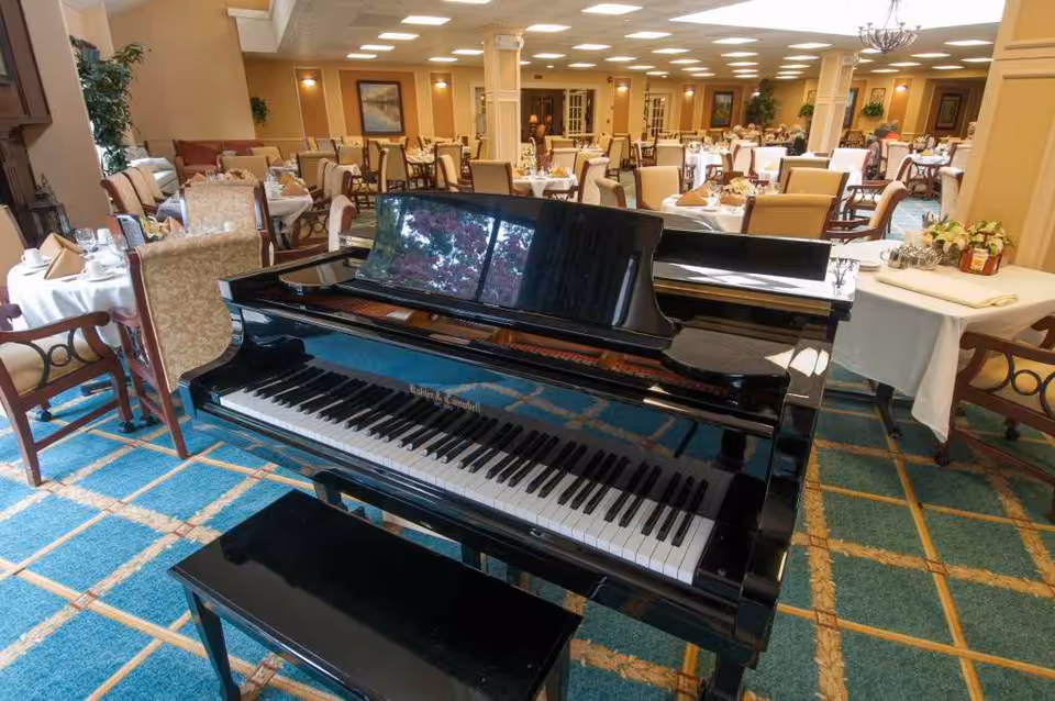 A black grand piano sits in the center of a large dining room with tables and chairs set for a meal.