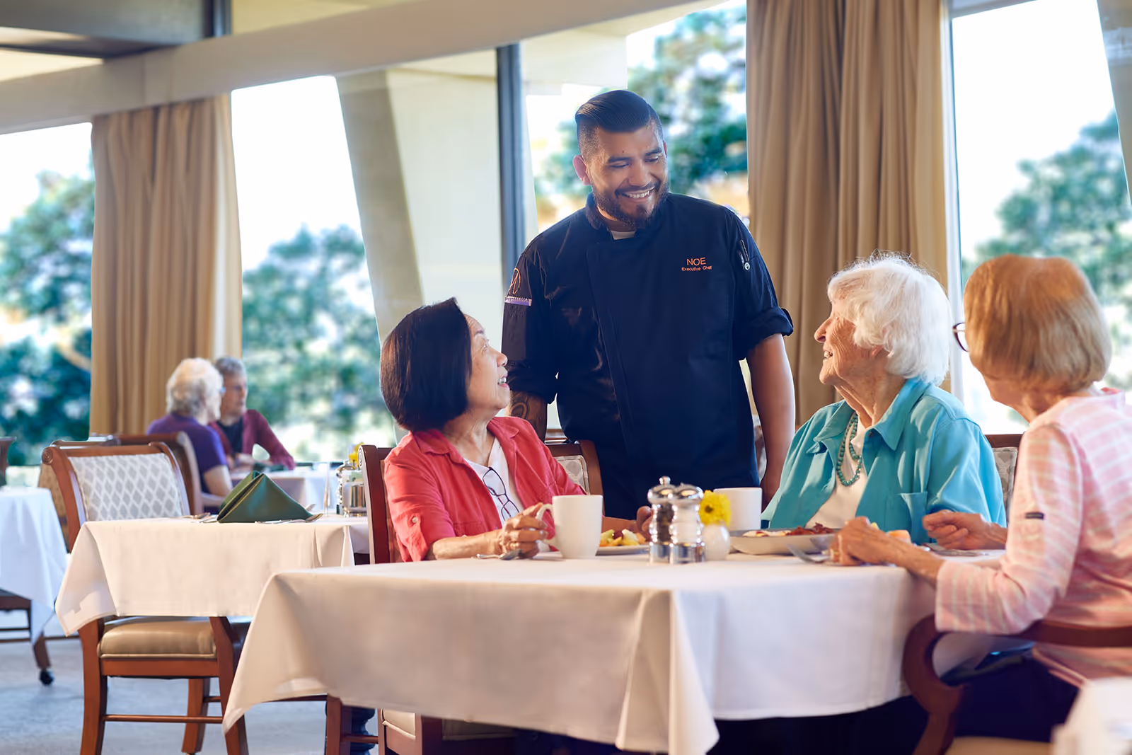 A chef in a black uniform smiling and talking to three elderly women seated at a dining table in a bright dining room with large windows and beige curtains. The table is set with white tablecloths, cups, plates, and a small flower vase.