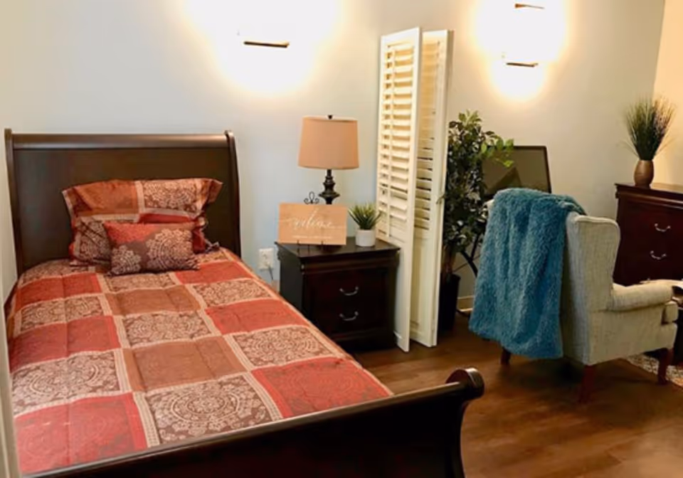 A cozy bedroom with a wooden bed featuring a red and brown patterned quilt and matching pillows. Next to the bed is a dark wooden nightstand with a lamp, a small plant, and a decorative sign. There is a white folding screen behind the nightstand, a green plant, a beige armchair with a blue throw draped over it, and a dark wooden dresser with another plant on top. The room has wooden flooring and soft wall lighting.