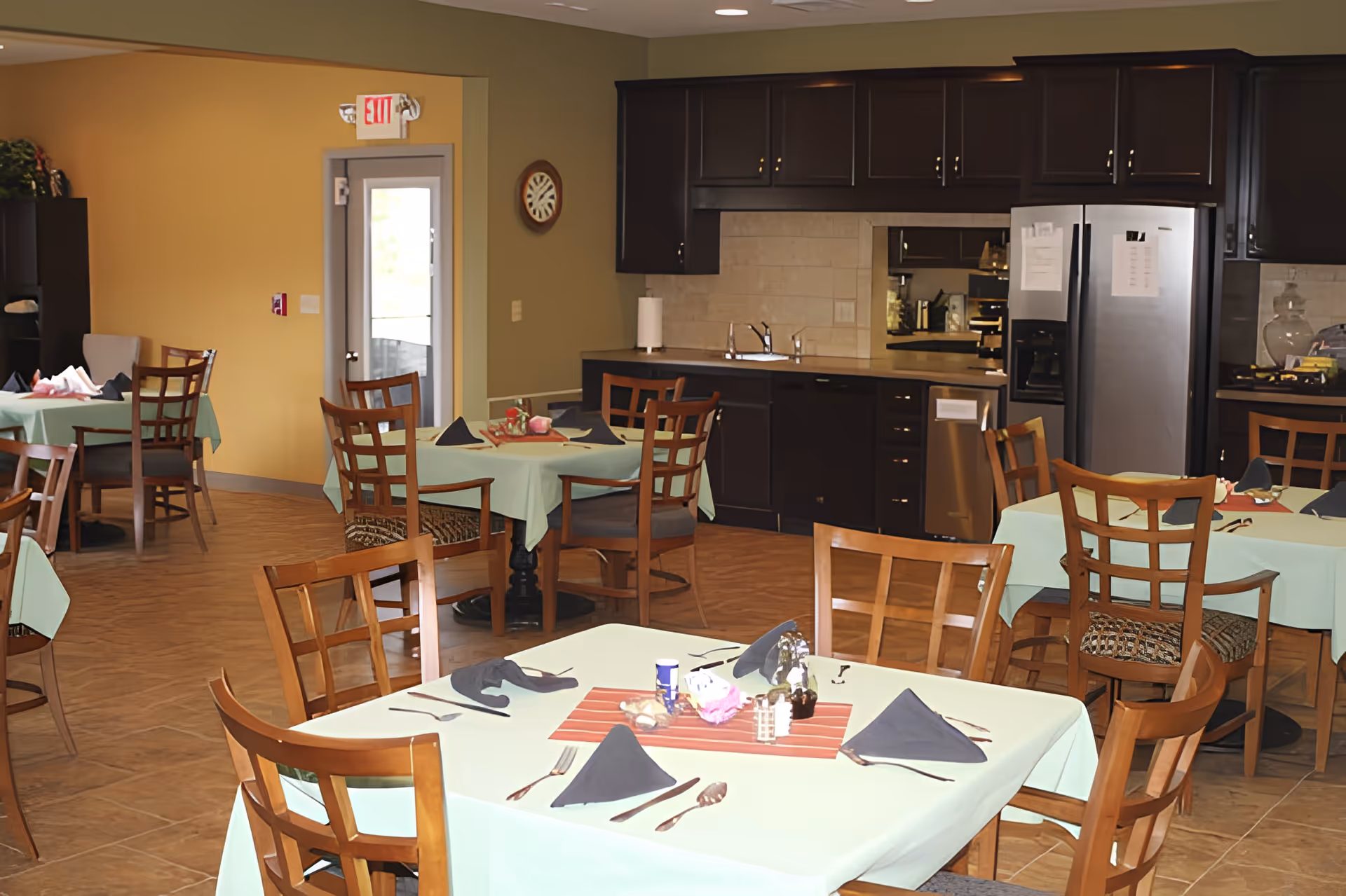 Dining room with multiple set tables and wooden chairs in front of a kitchen area with dark cabinets and a stainless refrigerator.