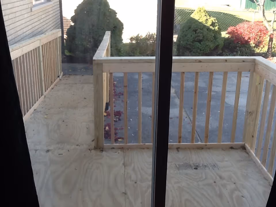 View through a glass door showing a small wooden balcony or porch with unfinished plywood flooring and wooden railings. Outside the balcony, there is a paved area and some greenery including bushes and trees with autumn-colored leaves.