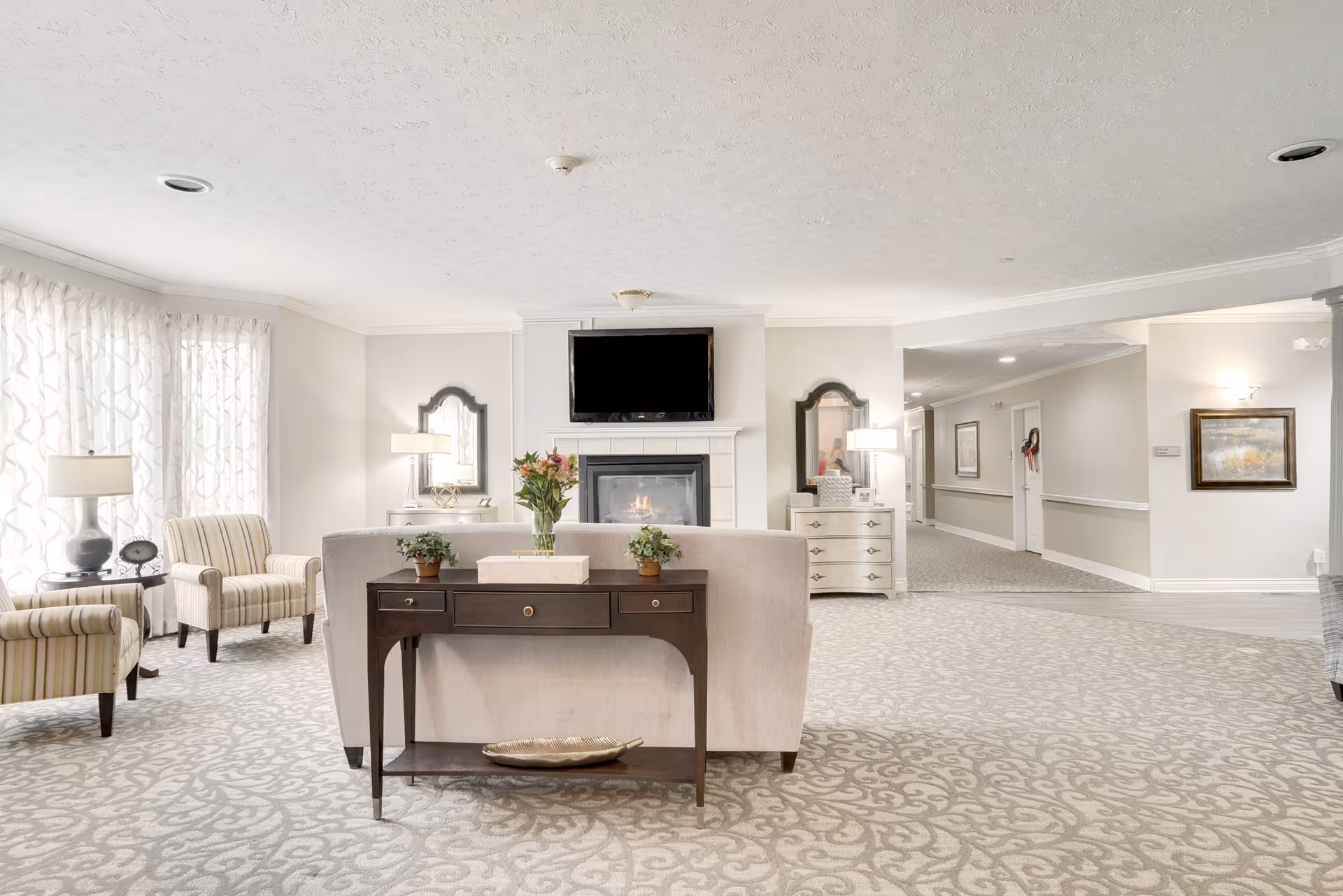 A bright and spacious living room area with a beige sofa facing a fireplace topped with a flat-screen TV. Two striped armchairs and a side table with a lamp are positioned near large windows with sheer curtains. The room features light-colored walls, patterned carpet, two mirrors above white dressers with lamps, and a hallway leading to other rooms.