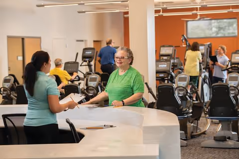 A senior woman in a green shirt smiling and talking to a staff member at a reception desk inside a fitness center. Several people are using exercise machines in the background.