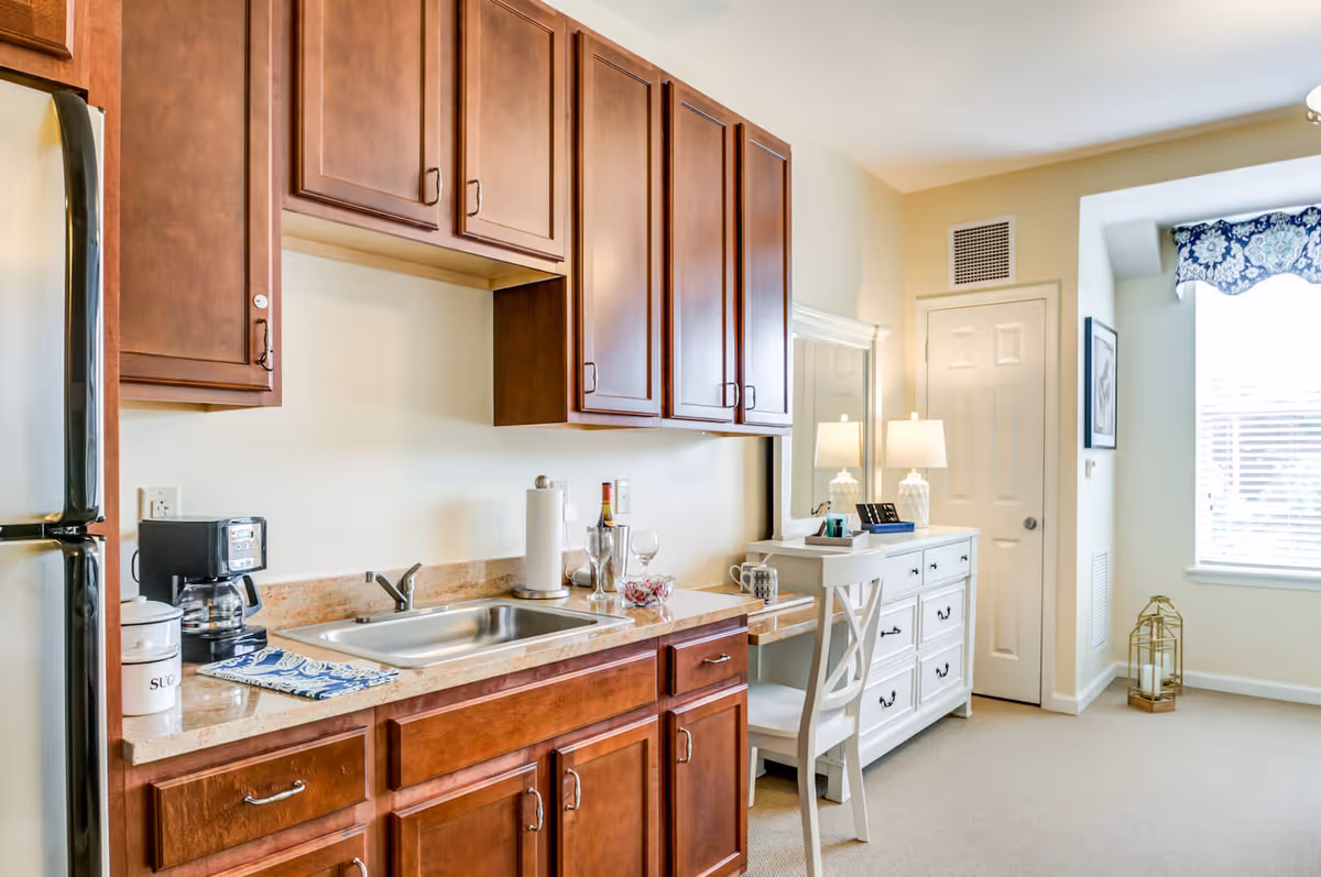 A bright kitchen area with wooden cabinets, a stainless steel sink, a coffee maker, and a refrigerator on the left. To the right, there is a white desk with a chair, two table lamps, and a dresser with drawers. A window with blinds and a blue patterned valance lets in natural light.