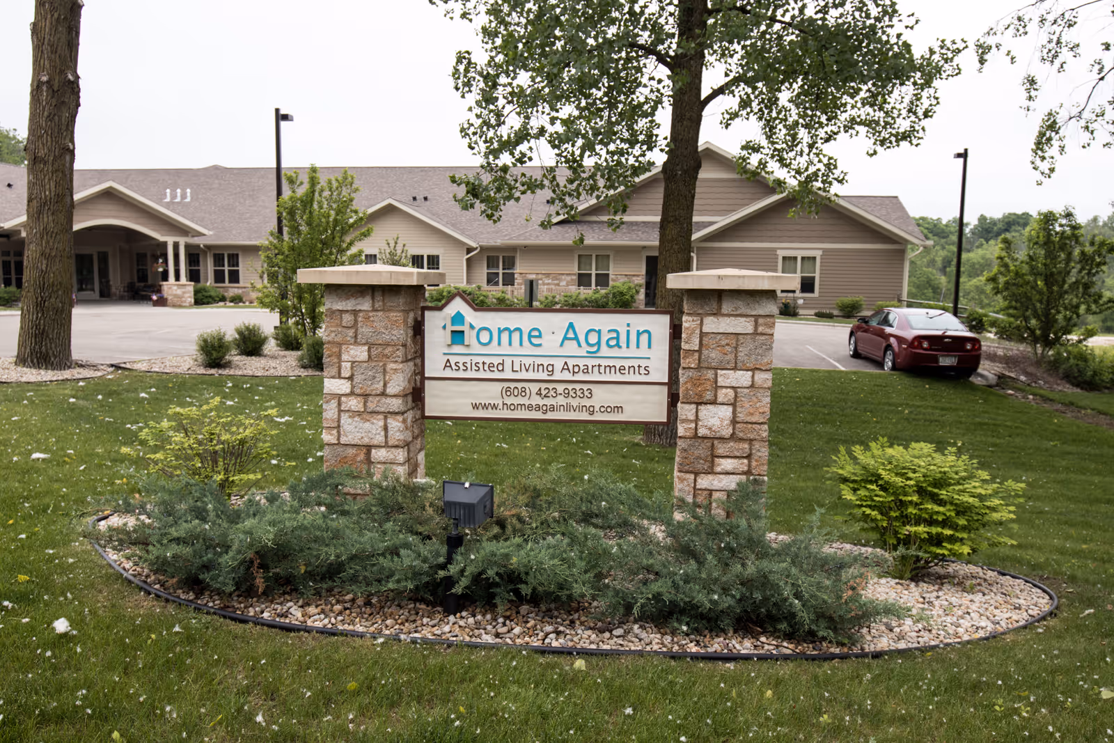 A stone sign reading "Home Again Assisted Living Apartments" on a landscaped lawn in front of a single-story assisted living building.