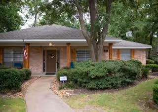 Front exterior of a single-story brick cottage with a walkway, porch columns, an American flag, and landscaped shrubs.