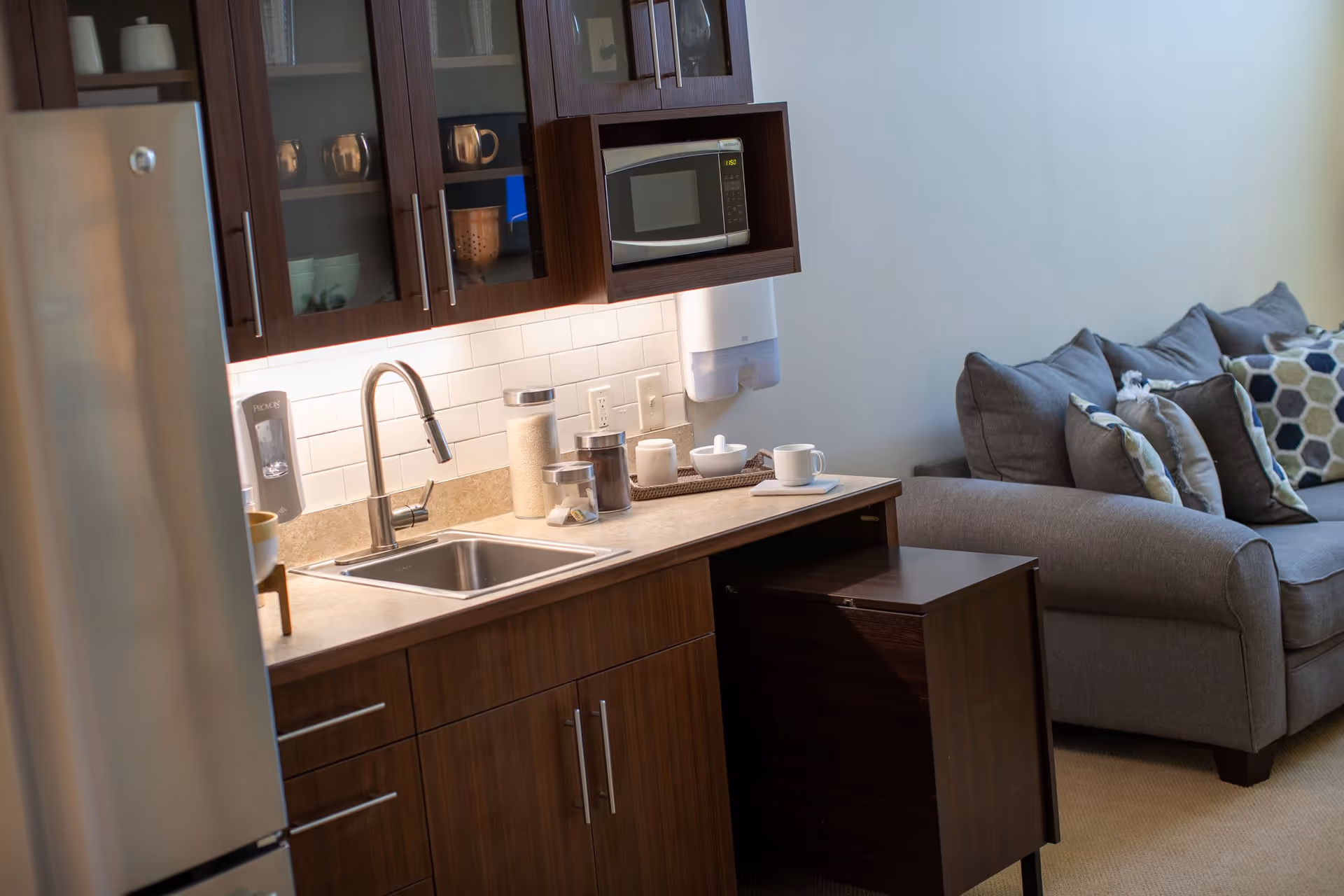A modern kitchenette with dark wood cabinets, a stainless steel sink, a microwave, and various jars on the countertop. To the right, there is a gray couch with multiple decorative pillows against a plain light-colored wall.