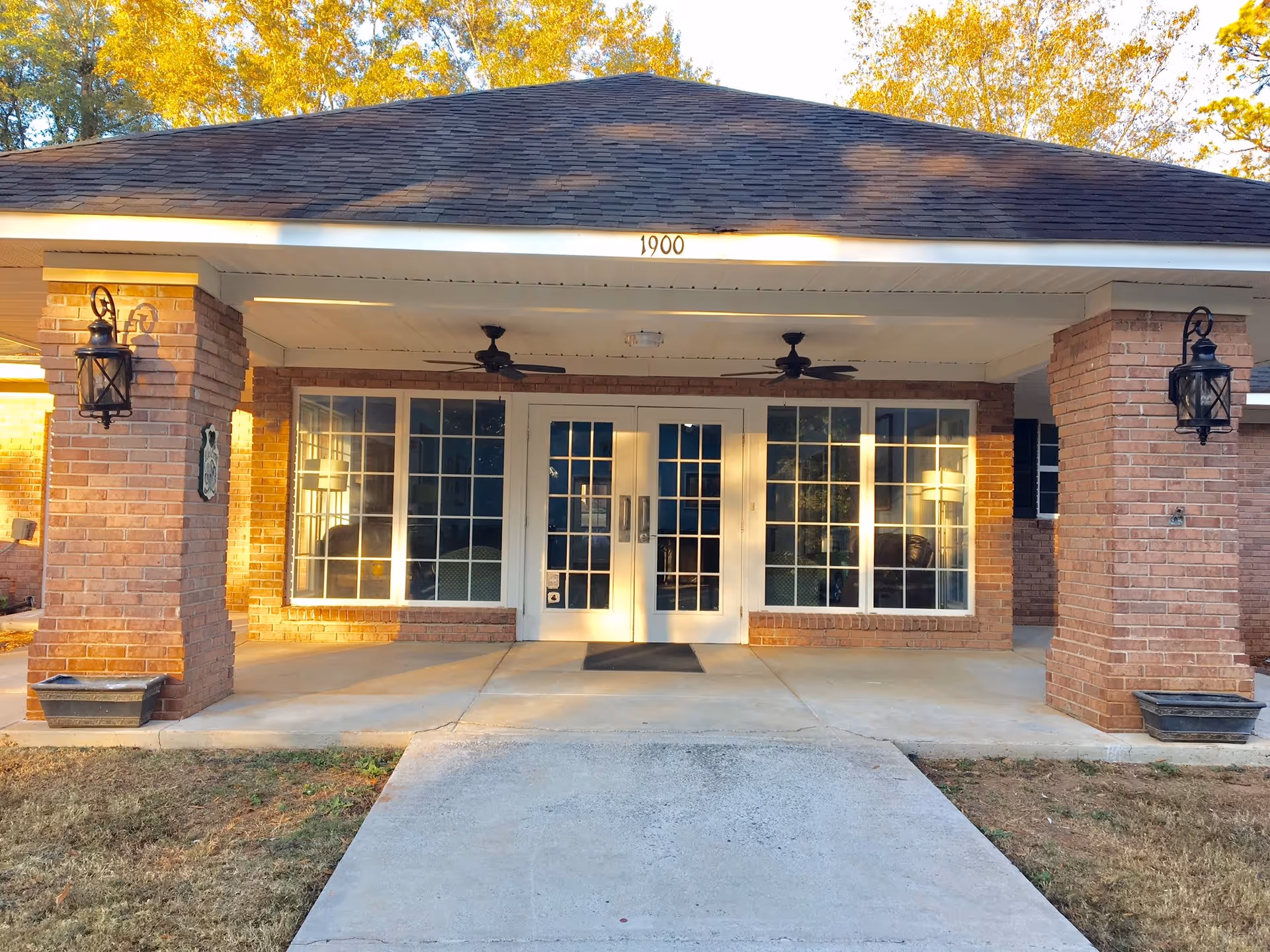 Front entrance of a brick building with large windows and double glass doors under a covered porch supported by brick columns. The address number 1900 is displayed above the entrance. Two black lantern-style wall lights are mounted on the columns, and ceiling fans are installed under the porch roof. Trees with yellow leaves are visible in the background.