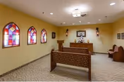 Interior of a small chapel or meditation room with beige walls, three stained glass windows on the left wall, wooden pews arranged in rows, a small altar with religious items, and soft overhead lighting.