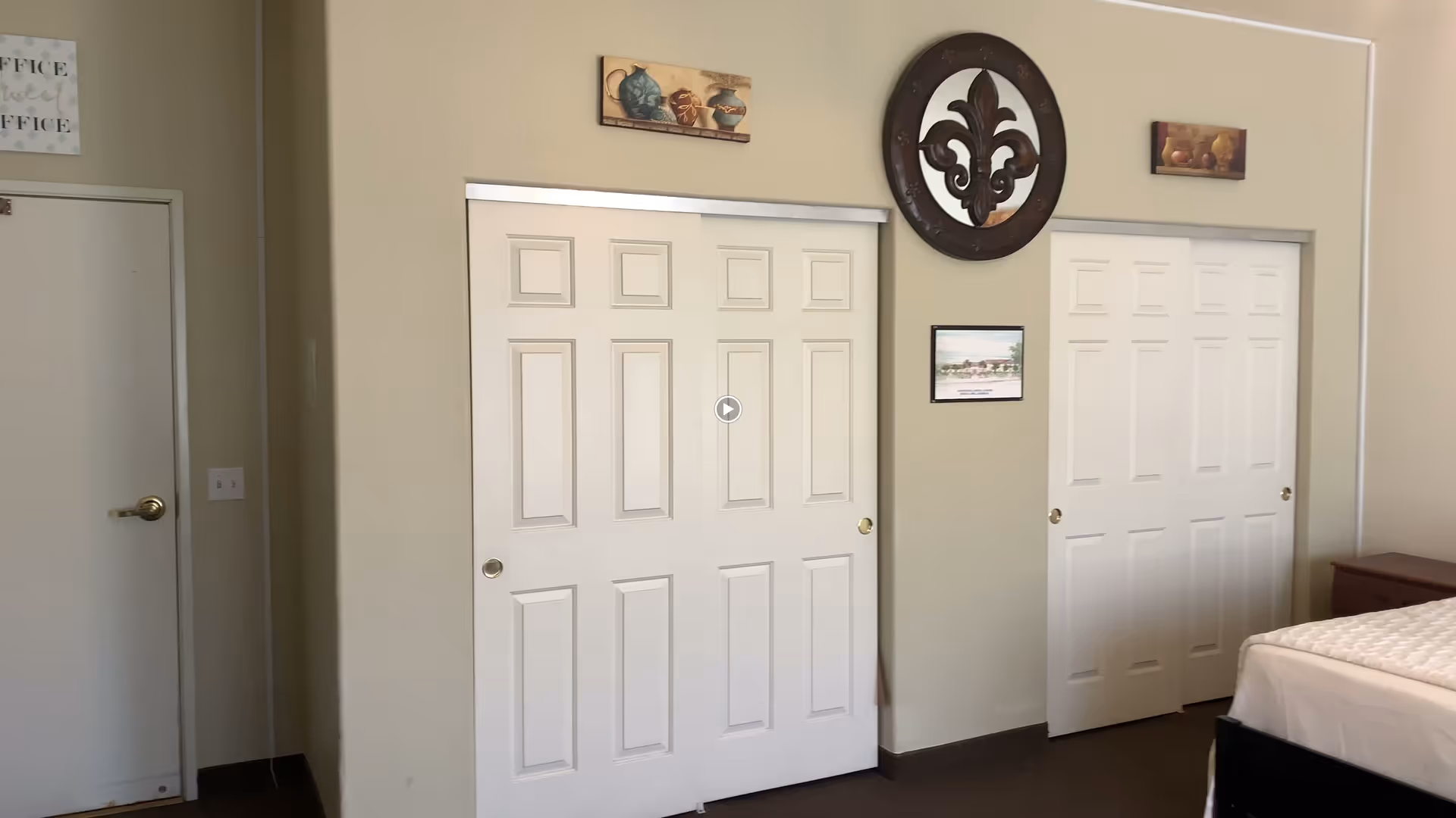 Interior view of a bedroom with two sets of white sliding closet doors, a bed with a white mattress on the right side, beige walls decorated with small framed pictures and a large circular wall ornament, and a closed white door with a brass handle on the left.
