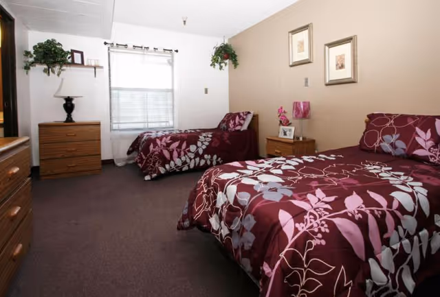 Furnished bedroom with two twin beds dressed in matching burgundy floral bedding, a nightstand, dressers, and a window.