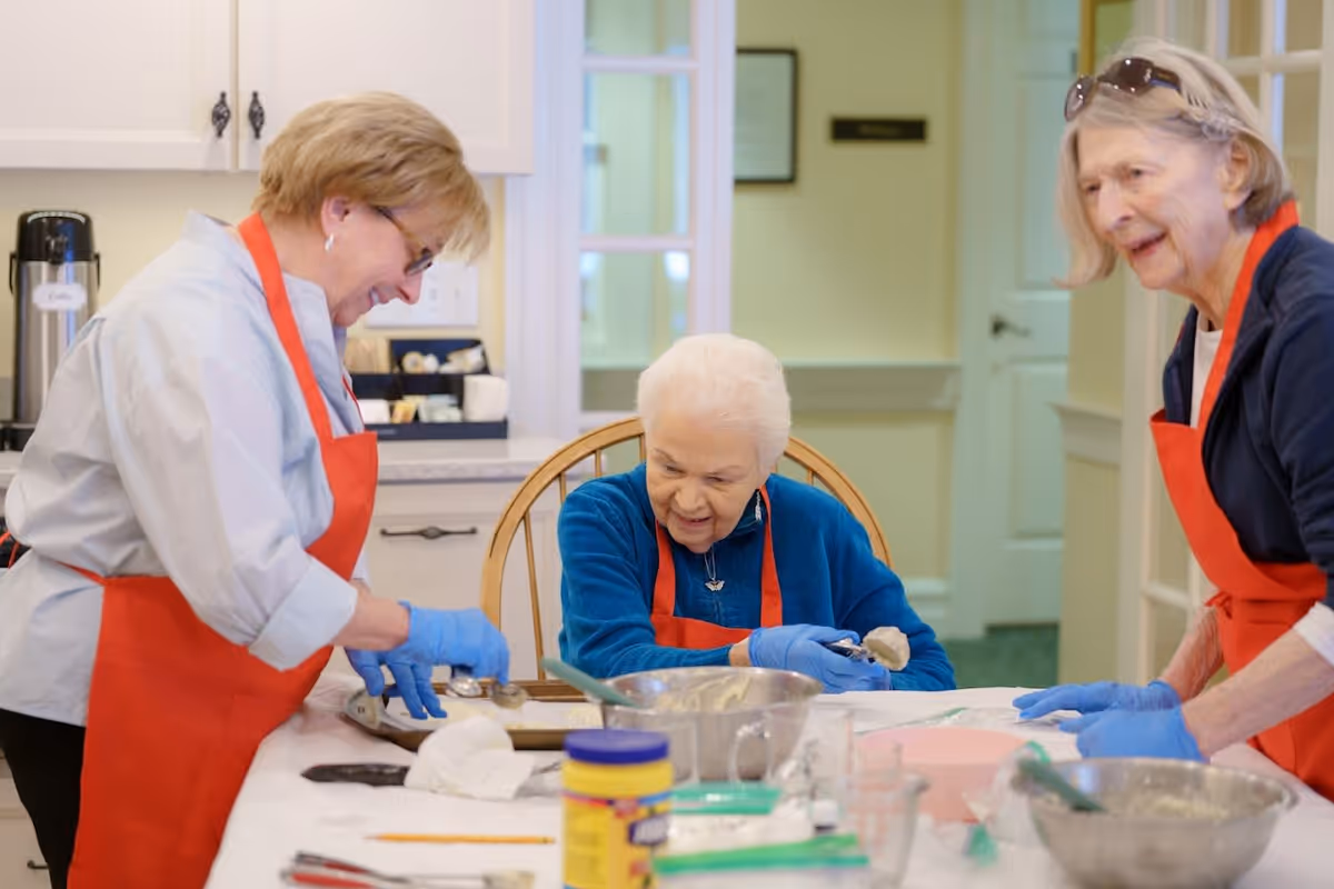 Three elderly women wearing red aprons and blue gloves are gathered around a kitchen table, engaging in a baking activity. One woman is seated and holding a scoop of dough, while the other two women stand on either side, smiling and assisting. Various baking utensils and ingredients are spread out on the table.