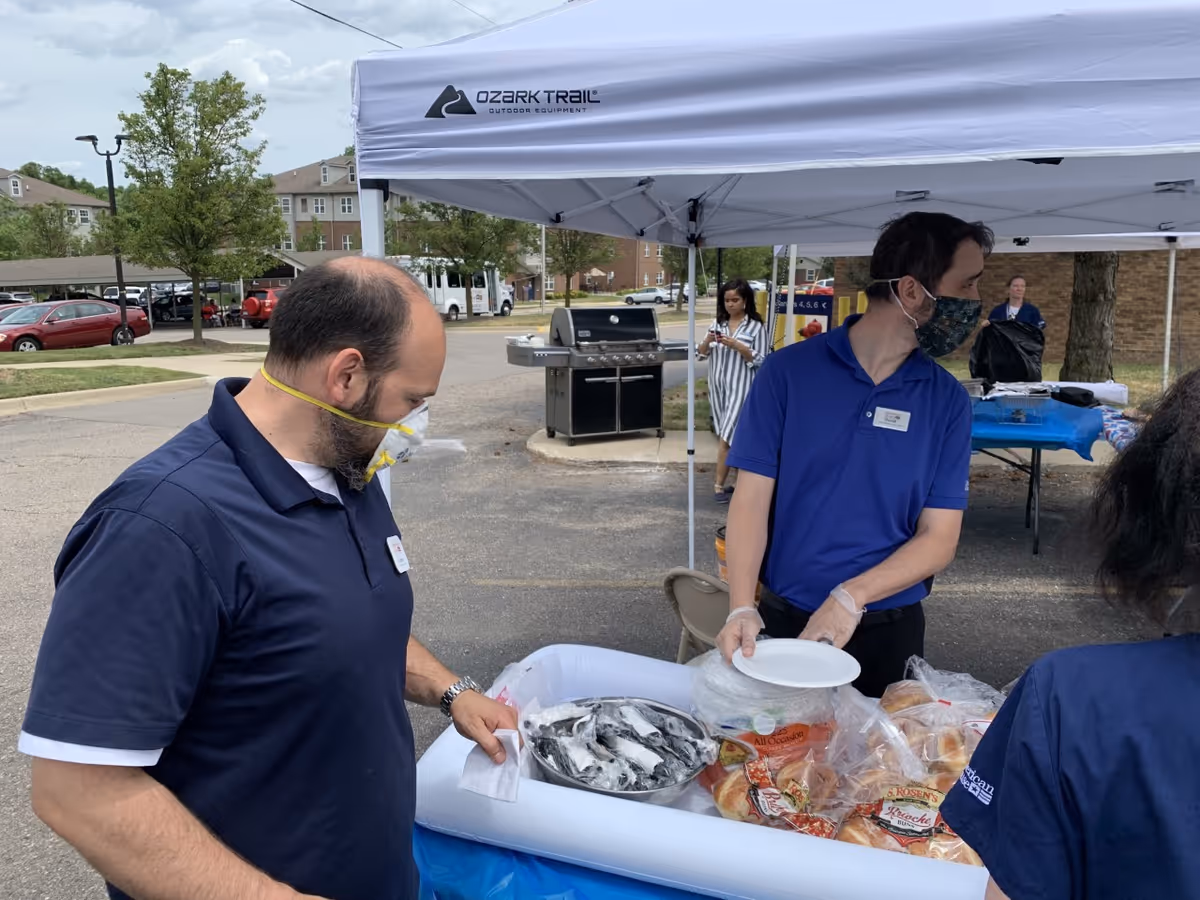 Two men wearing masks and gloves stand under a white Ozark Trail canopy tent at an outdoor event. One man is holding a stack of white plates while the other is looking down at a table filled with bread rolls and a bowl of ice. In the background, a woman in a striped dress is using her phone, and there are parked cars and buildings visible.