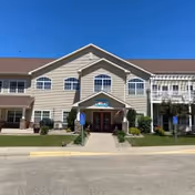 Two-story beige senior living building with a central covered entrance, columns, landscaped shrubs, and a clear blue sky.
