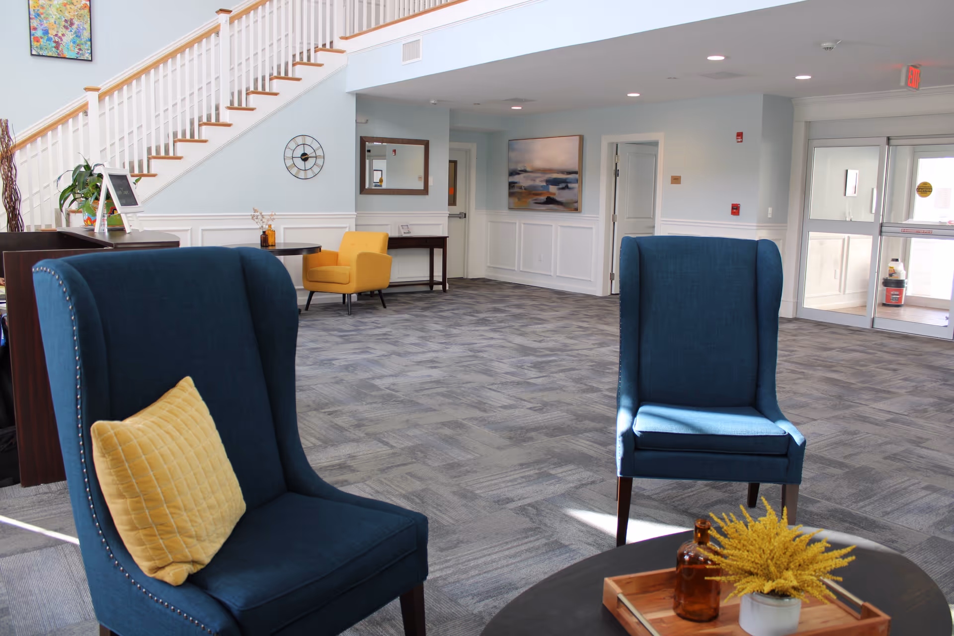 A bright and spacious senior living facility lobby with two blue armchairs featuring yellow pillows, a round table with decorative items, a yellow armchair in the background, a staircase with white railing, wall art, a clock, and a glass entrance door.