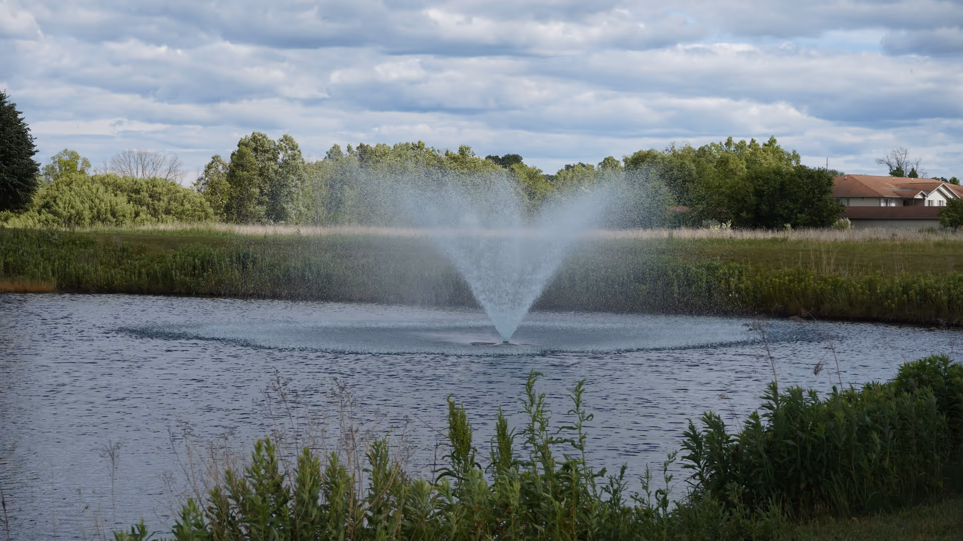A pond with a central fountain spraying water surrounded by grassy banks, trees, and distant houses under a cloudy sky.
