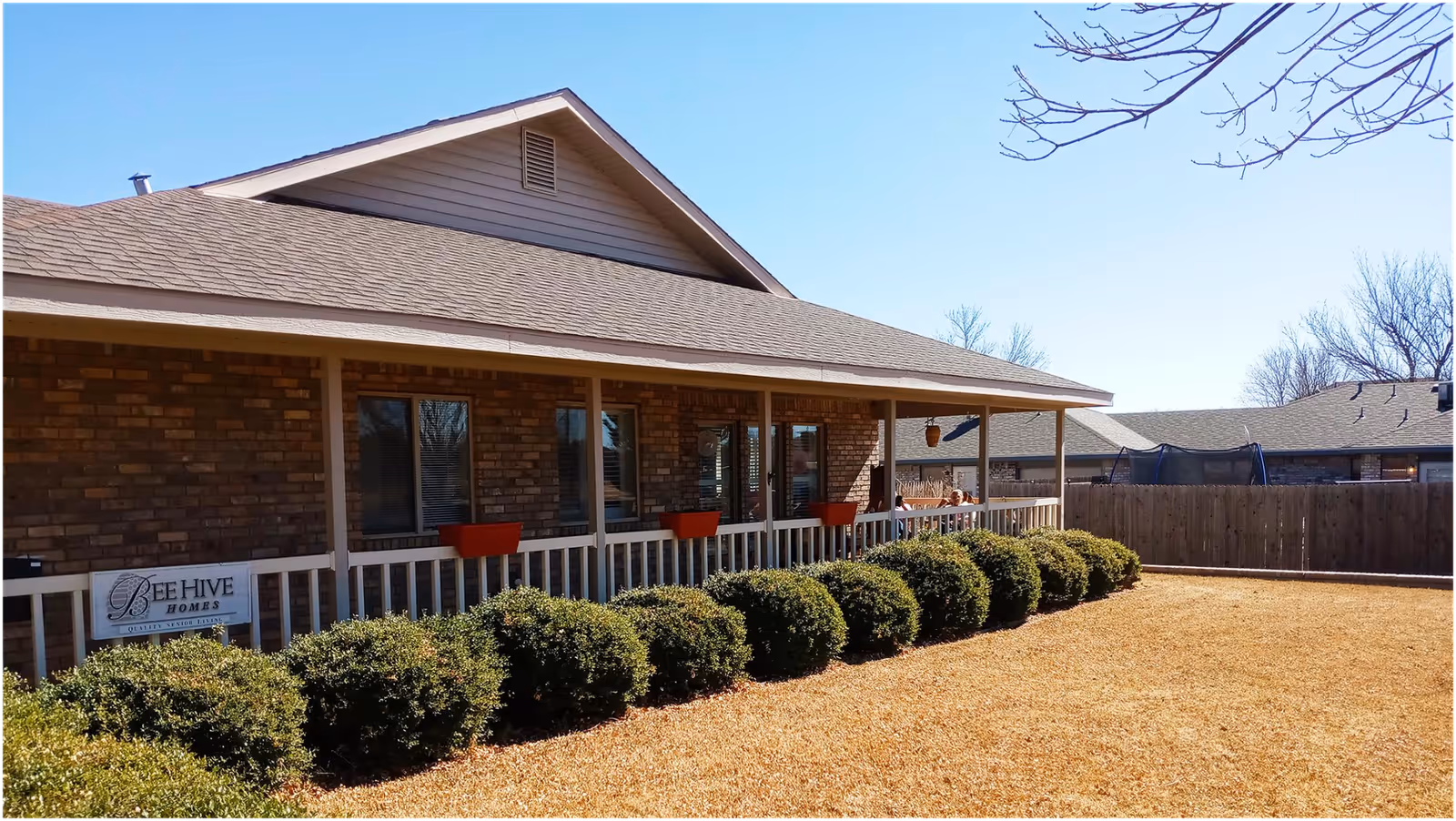 Exterior view of a single-story brick building with a covered porch and white railing. There are several windows along the building and a row of neatly trimmed bushes in front. The sky is clear and blue, and a wooden fence encloses the yard area. A sign on the porch reads 'BeeHive Homes'.