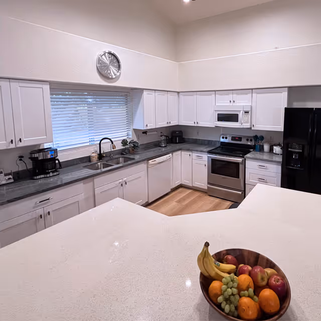 A modern kitchen with white cabinets, gray countertops, and stainless steel appliances including an oven and microwave. There is a large white island countertop in the foreground with a wooden bowl containing bananas, grapes, apples, and oranges. A window with white blinds and a wall clock is above the sink area.