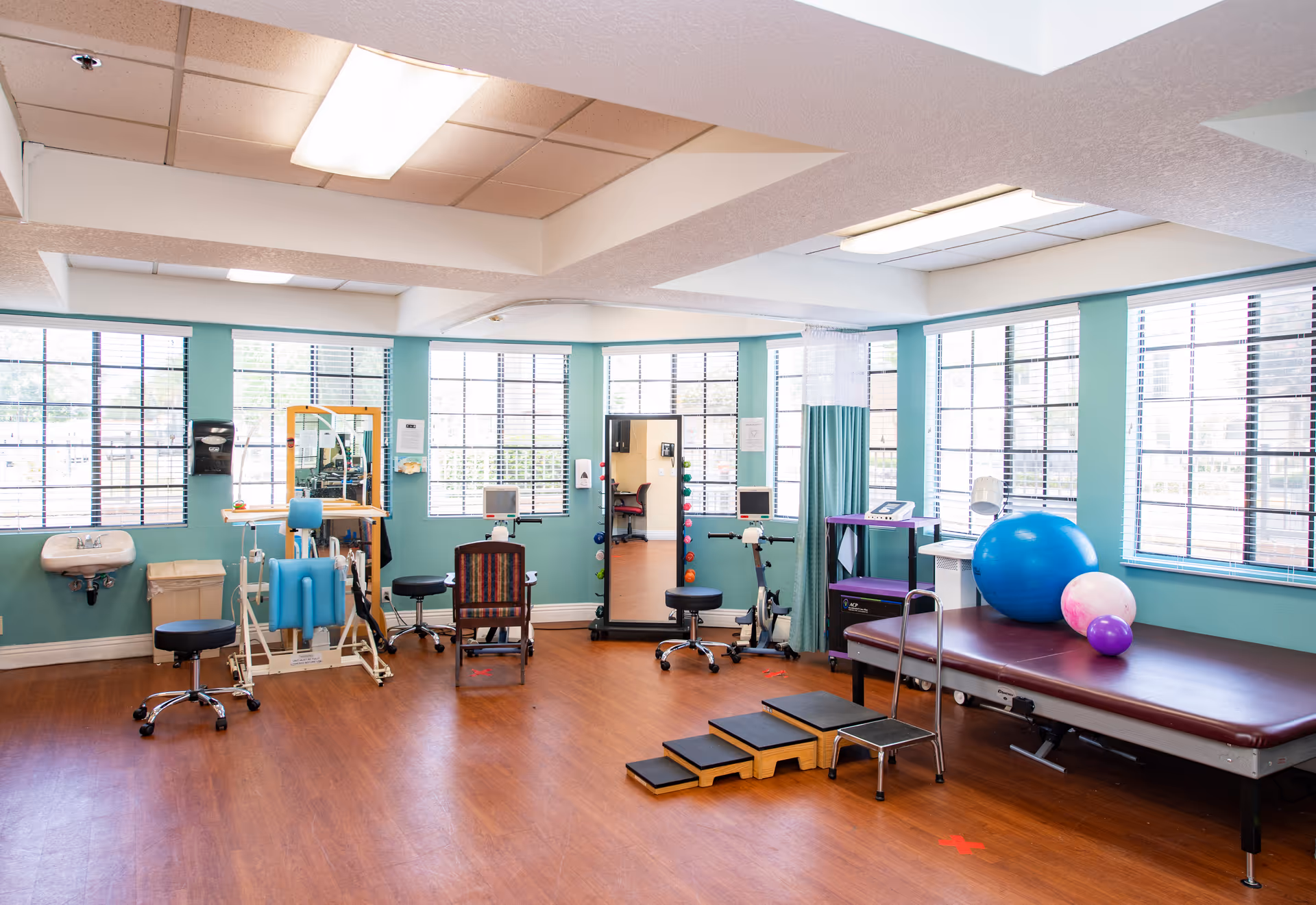 Bright rehabilitation/exercise room with therapy equipment, treatment table, exercise balls, and large windows.