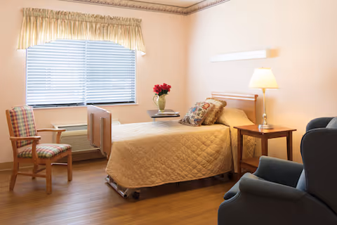 A cozy bedroom with a single bed covered in a beige quilt, a wooden side table with a lamp, a small table with a vase of red flowers, a plaid upholstered chair, and a blue armchair. The room has a window with blinds and a valance, light-colored walls, and wood flooring.