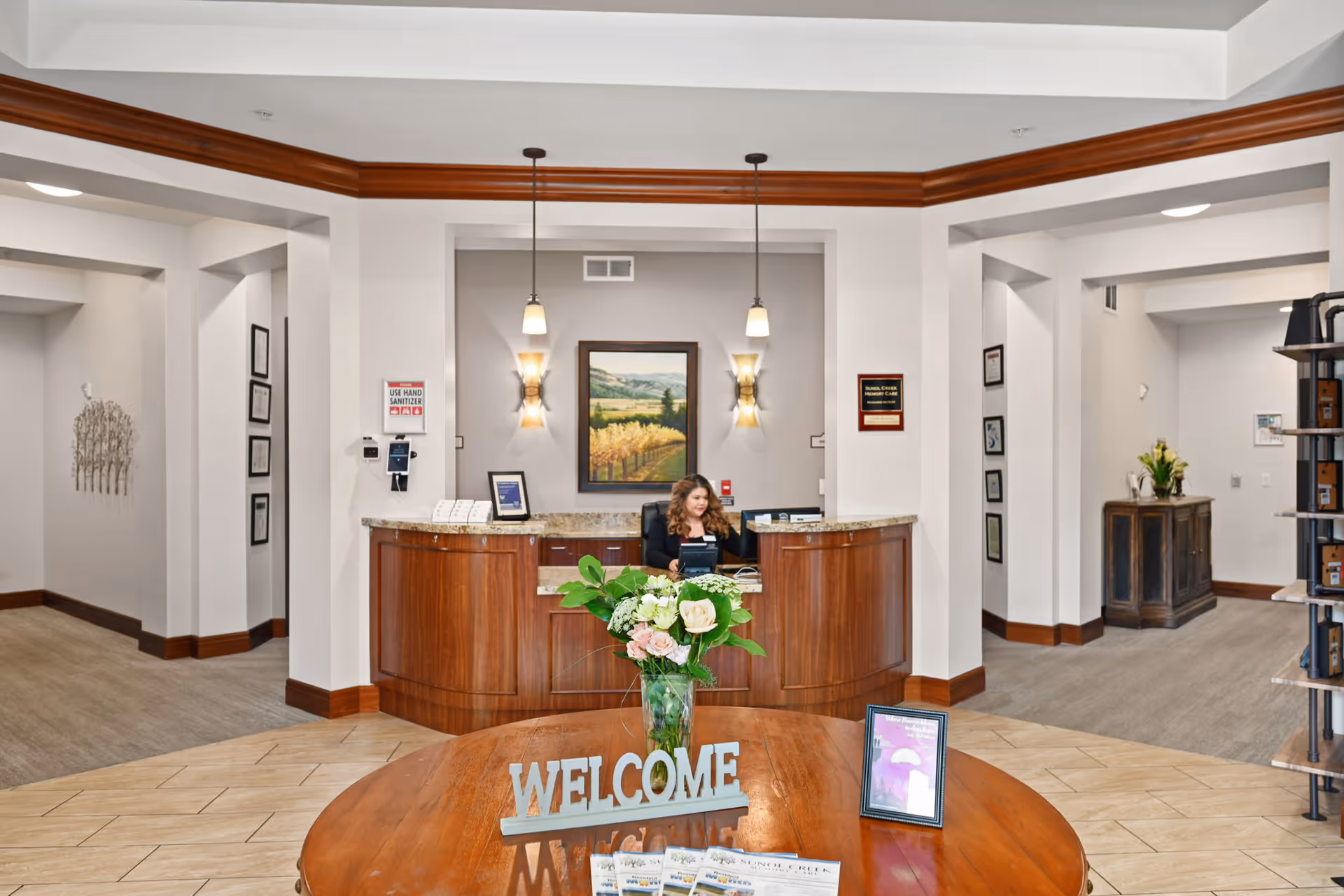 Reception area of Sunol Creek Memory Care facility with a wooden front desk where a woman is seated. The area features a round wooden table with a vase of flowers and a 'WELCOME' sign. The walls are white with wooden trim, and there is a framed landscape painting behind the desk. Two pendant lights hang from the ceiling above the desk.