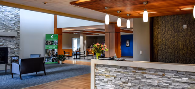 Interior view of a senior living facility lobby area with a stone and wood reception desk in the foreground, modern pendant lights hanging from a wooden ceiling, a seating area with chairs near a stone fireplace, and a green informational banner in the background.
