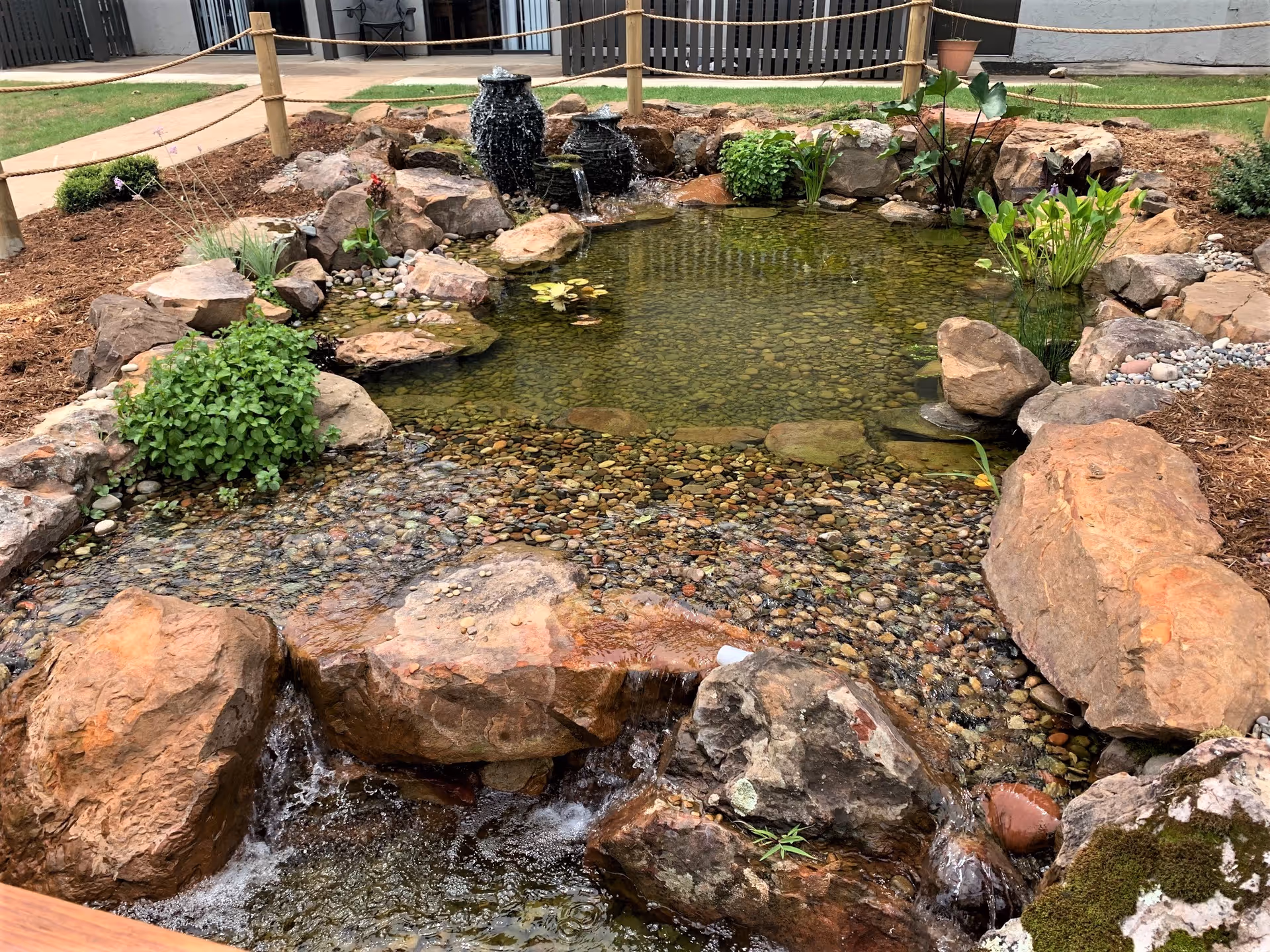 Rock-lined pond with a waterfall, surrounded by rocks and garden near a building.