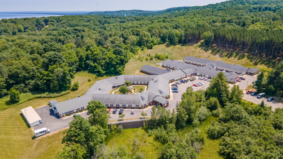 Aerial view of Brookridge Heights facility surrounded by dense green forest and trees, with a large parking area and multiple connected buildings forming a U-shape. The landscape includes grassy areas and a distant view of a body of water on the horizon.