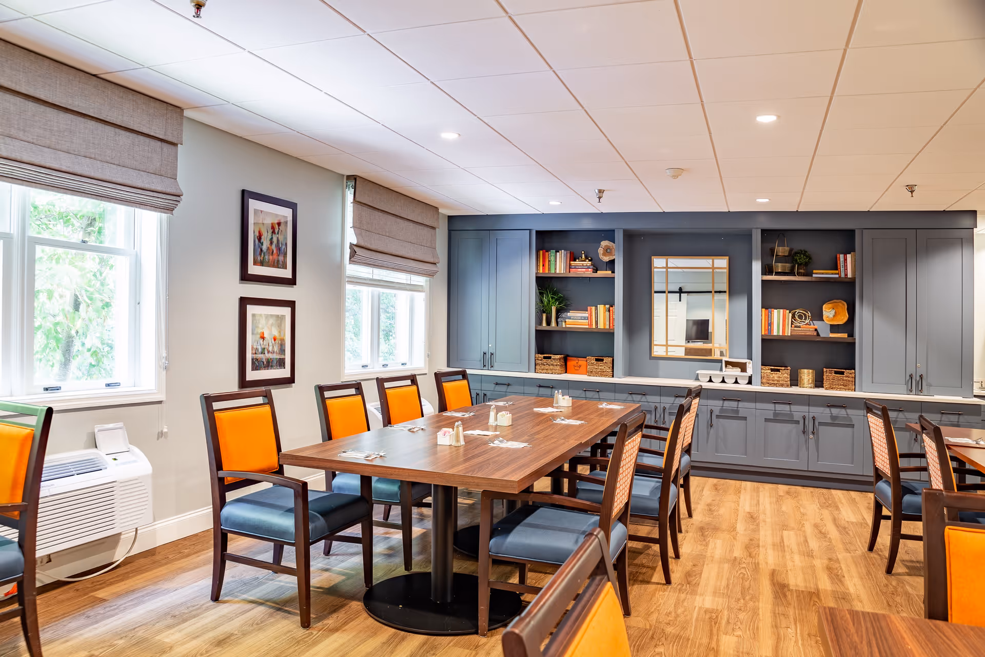 A bright dining room with wooden tables and chairs featuring orange and blue cushions. The room has large windows with beige Roman shades, light wood flooring, and a built-in gray cabinet with shelves holding books, baskets, and decorative items. The ceiling has recessed lighting.