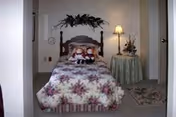 A small bedroom with a single bed covered in a floral bedspread. Two dolls are placed on the bed. There is a decorative floral arrangement hanging above the headboard. To the right of the bed is a round table with a green tablecloth, a lamp, and a floral arrangement. A small rug is on the floor near the table.