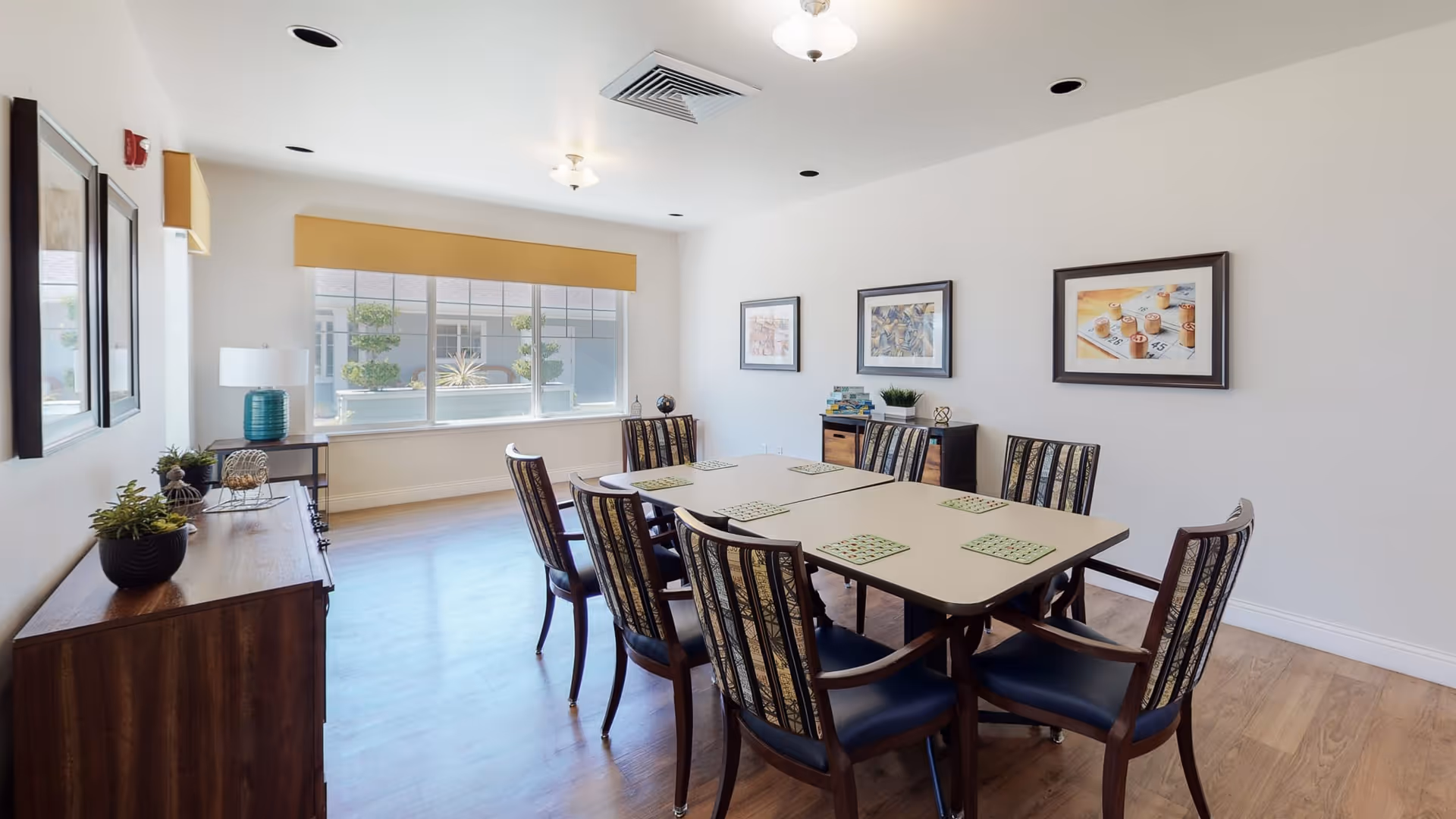 A bright and clean dining room with a rectangular table surrounded by eight chairs. The table has green placemats, and the room features wooden flooring, white walls, framed artwork, and a large window with a yellow valance letting in natural light. There are decorative plants and a lamp on wooden sideboards along the walls.