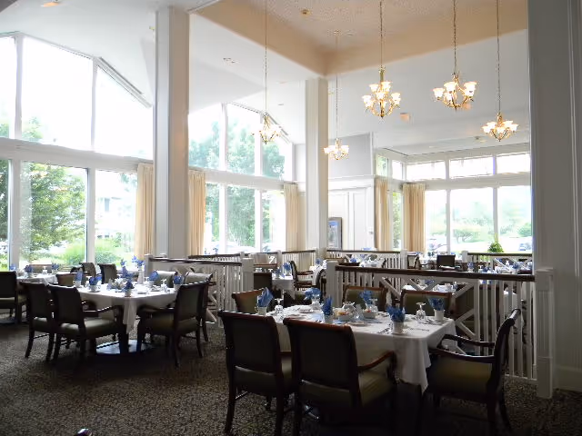 A bright dining room with large windows letting in natural light. Several tables are set with white tablecloths, blue napkins, and dining ware. The room features high ceilings with hanging chandeliers and a carpeted floor. Outside greenery is visible through the windows.