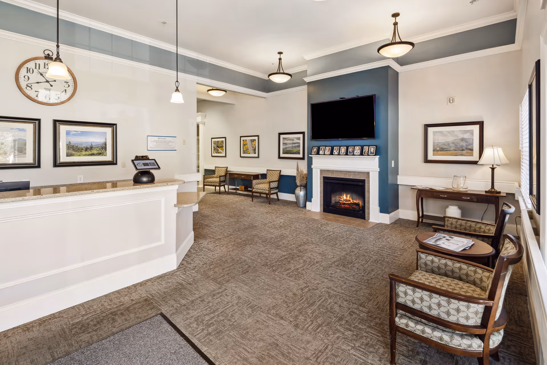 A spacious and well-lit reception area with a white front desk on the left, a large wall clock above it, and framed landscape pictures on the walls. The room features a cozy fireplace with a TV mounted above it, several chairs with patterned upholstery, small tables, and a lamp on a side table near the window. The carpeted floor and pendant lights add to the welcoming atmosphere.