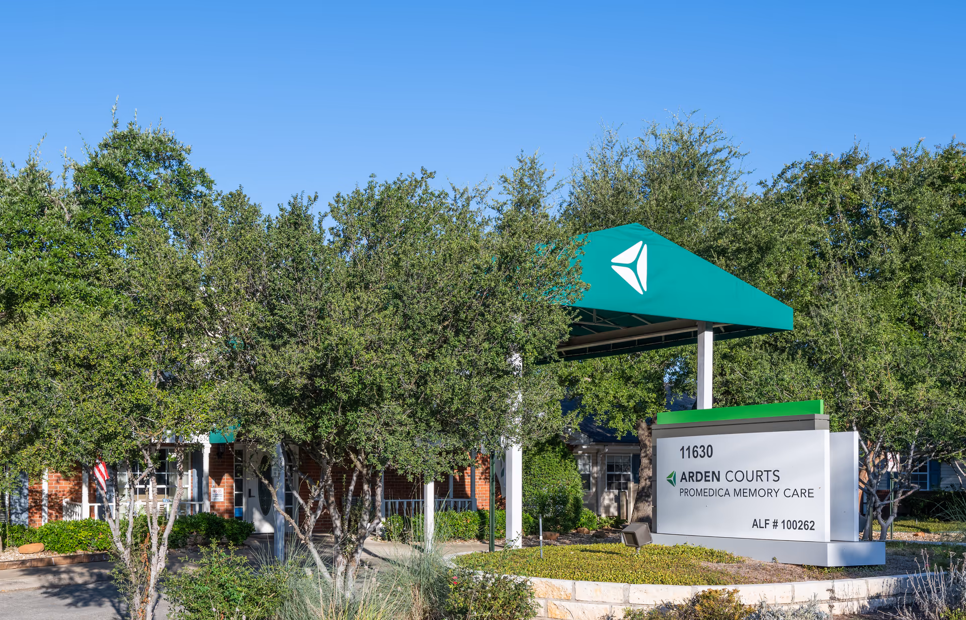 Entrance to Arden Courts ProMedica Memory Care with a green canopy and sign in front of a brick building surrounded by trees.
