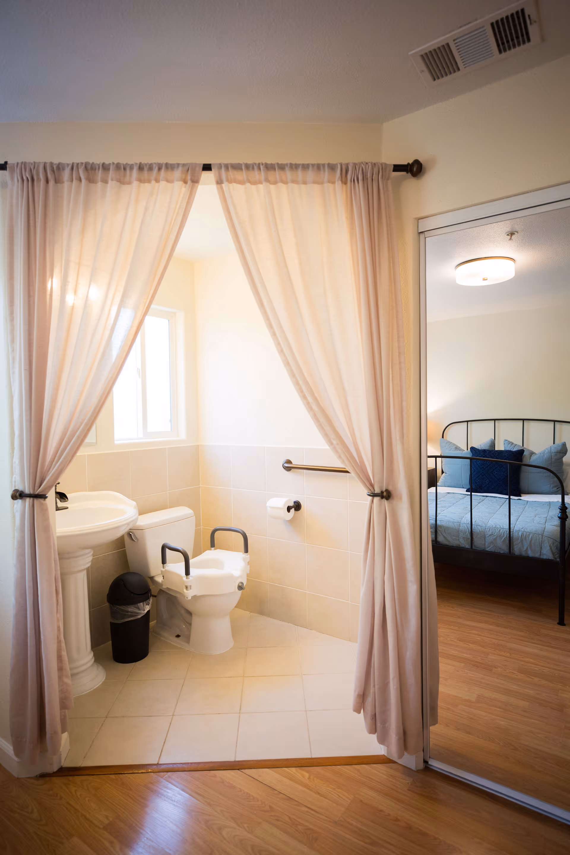 A small accessible bathroom with a raised toilet with handrails and a pedestal sink seen through parted curtains, with a bed visible in the adjacent room.