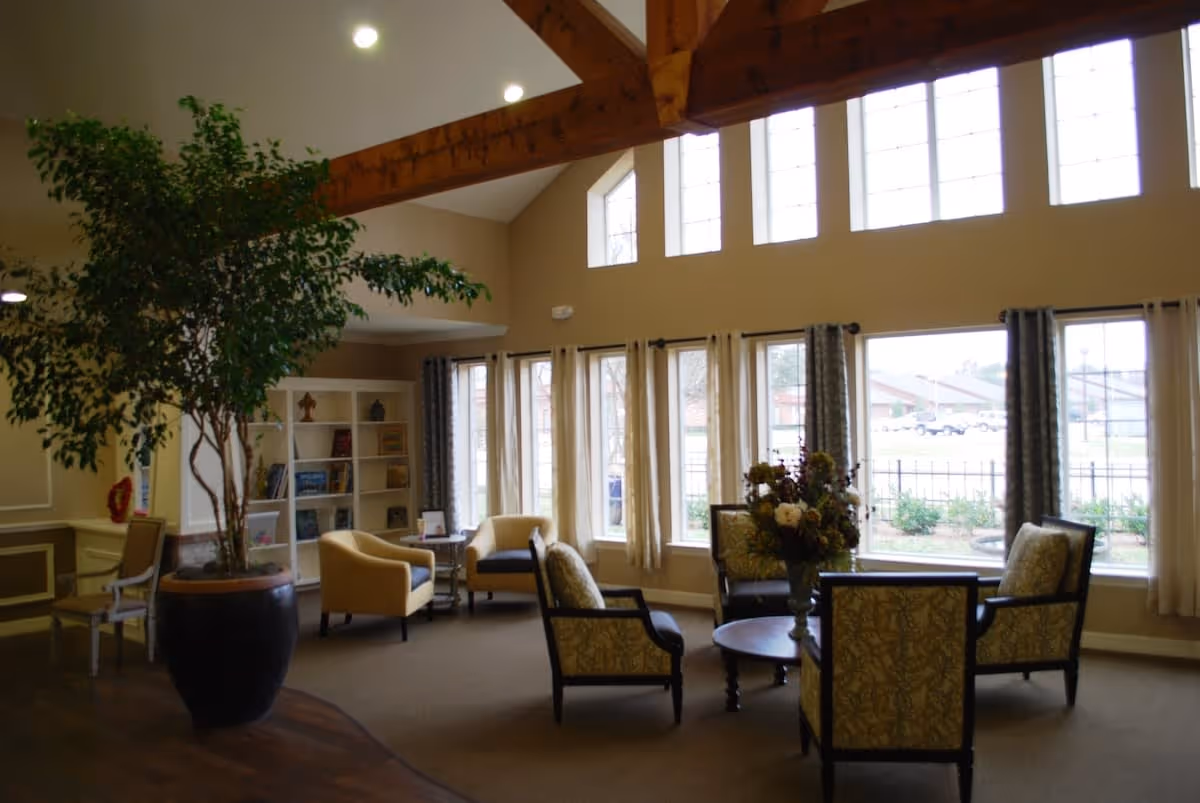 A spacious living room area with large windows letting in natural light. The room features a high ceiling with exposed wooden beams, several armchairs arranged around a round coffee table with a floral centerpiece, a large potted plant, and a bookshelf filled with books and decorative items. Light curtains frame the windows, and the floor is a combination of carpet and wood.