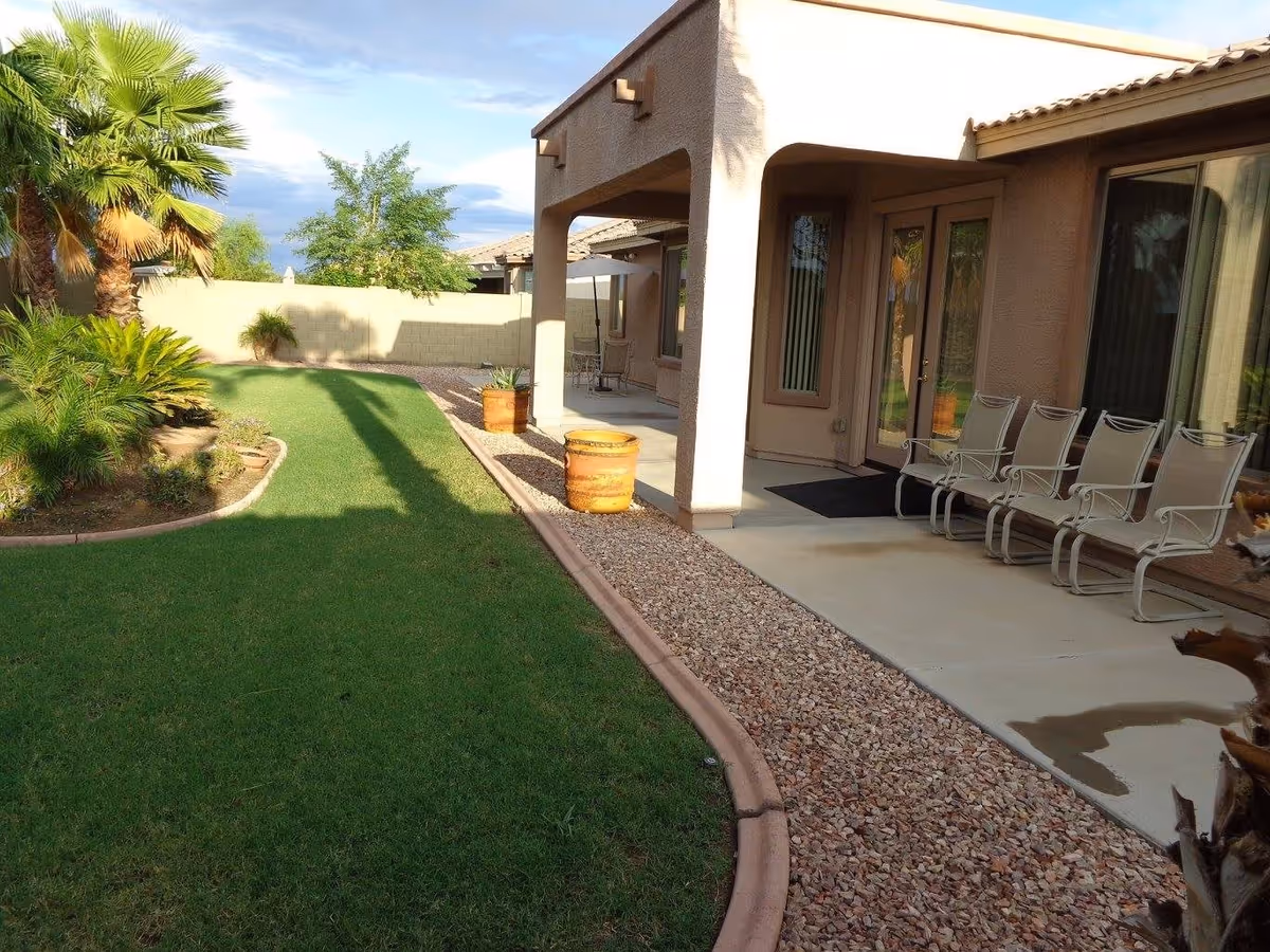Outdoor patio area of a residential facility with a covered porch featuring four metal chairs, potted plants, a well-maintained green lawn, palm trees, and a stone border along the walkway.