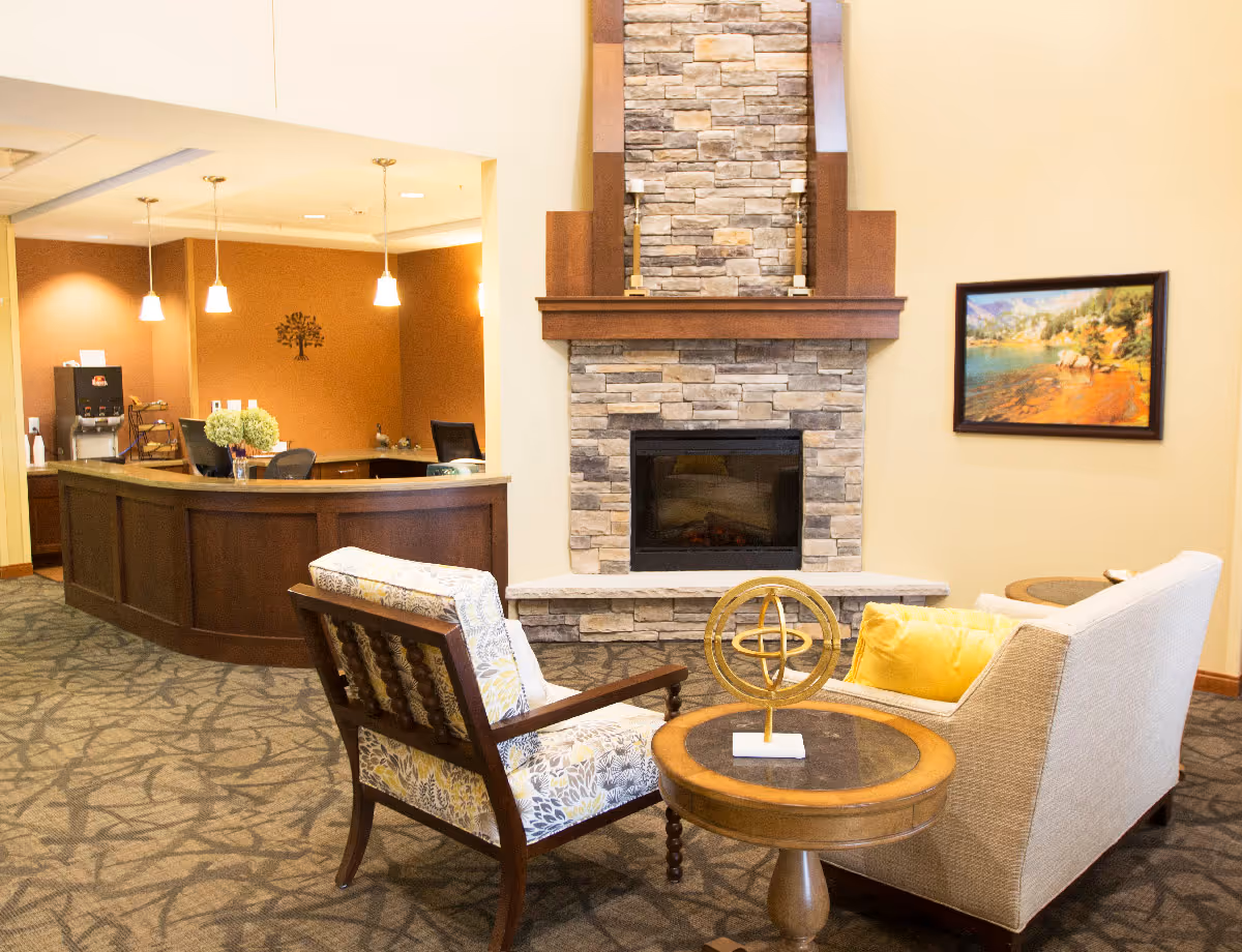 Comfortable lobby area with chairs and a round table facing a stone fireplace and a reception desk in the background.