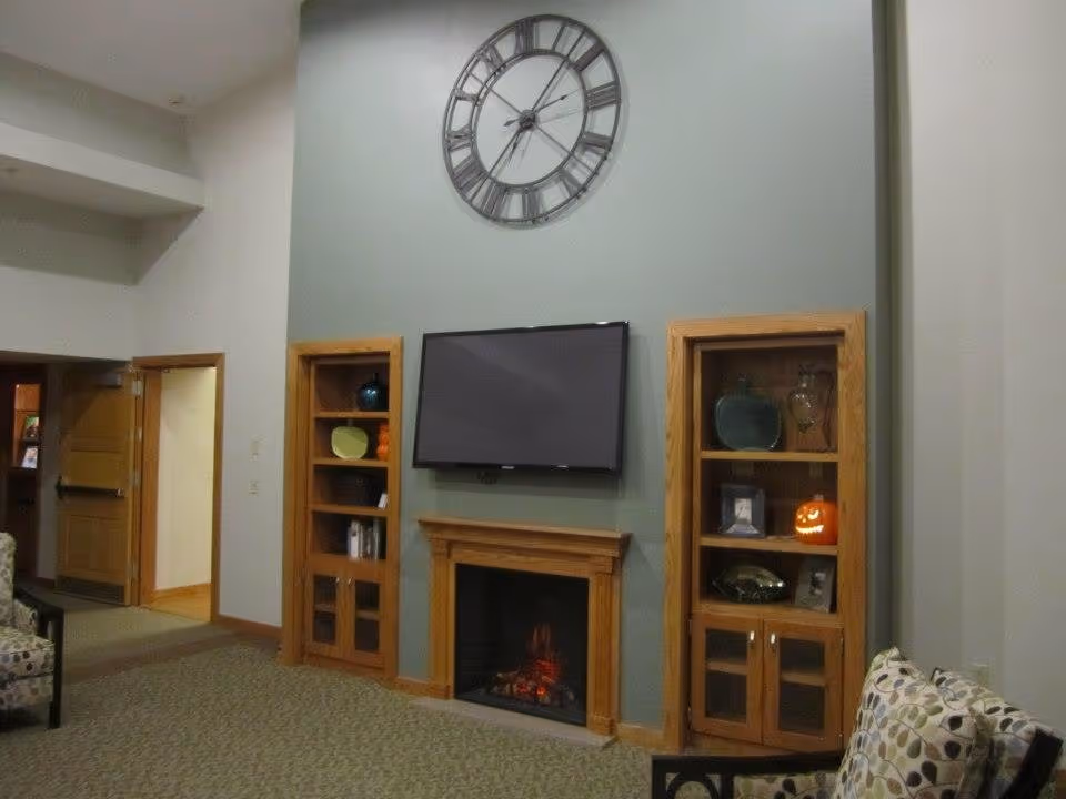 A cozy living room area with a wall-mounted flat screen TV above a wooden fireplace. On either side of the fireplace are wooden built-in shelves with decorative items including a pumpkin decoration. Above the TV is a large wall clock with Roman numerals. The room has carpeted flooring and patterned cushioned chairs.