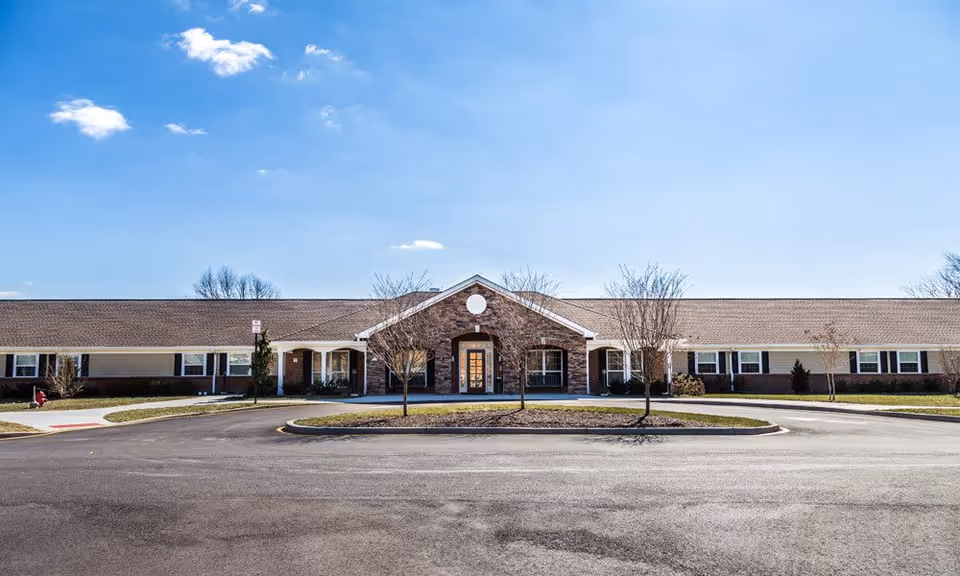 Front exterior view of a single-story senior living facility building with a peaked entrance, brick and siding facade, several windows, small trees, and a circular driveway under a blue sky with some clouds.